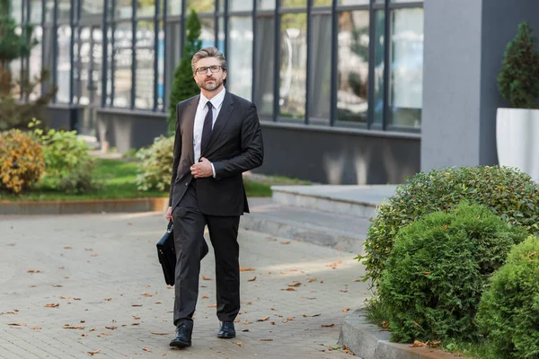 Schöner Diplomat mit Brille, Aktentasche in der Hand und zu Fuß in der Nähe des Gebäudes — Stockfoto