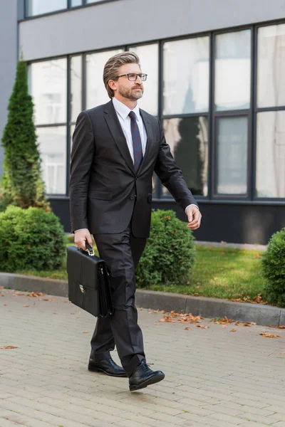 Bearded diplomat in glasses holding briefcase and walking near building — Stock Photo