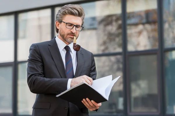 Handsome diplomat smoking pipe and looking at folder with documents — Stock Photo