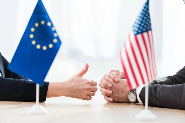 Vista cortada de diplomata da união europeia mostrando polegar para cima perto embaixador dos EUA sentado com as mãos apertadas — Fotografia de Stock