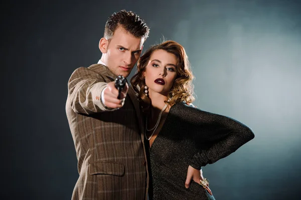 Selective focus of dangerous gangster holding gun and standing near attractive woman  on black — Stock Photo