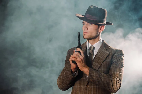 Handsome gangster in hat holding gun on black with smoke — Stock Photo