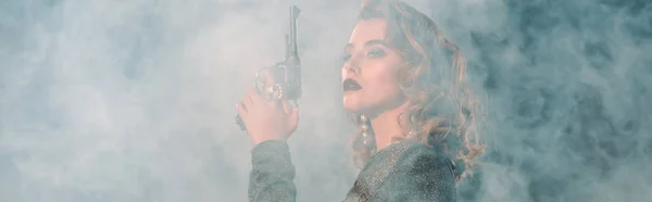 Tiro panorámico de mujer atractiva sosteniendo arma cerca del humo - foto de stock