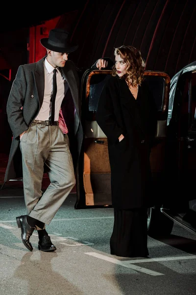 Красивый гангстер, стоящий с рукой в кармане и глядя на привлекательную женщину возле машины — стоковое фото
