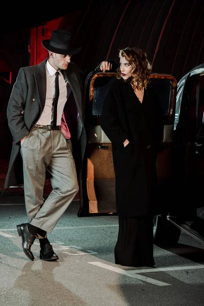 Handsome gangster standing with hand in pocket and looking at attractive woman near car — Stock Photo