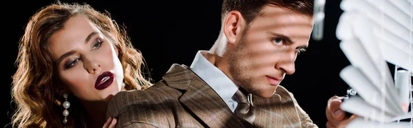 Panoramic shot of handsome man touching window blinds near attractive woman isolated on black — Stock Photo