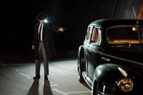 Handsome gangster in hat and coat holding gun near retro car — Stock Photo