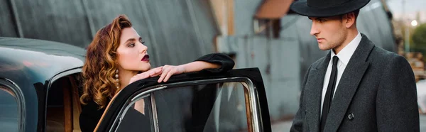 Panoramic shot of handsome man in hat standing near attractive woman in retro car — Stock Photo