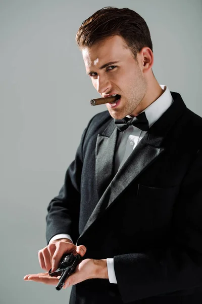 Elegante gángster en traje con corbata de lazo sosteniendo arma mientras fuma cigarro aislado en gris - foto de stock