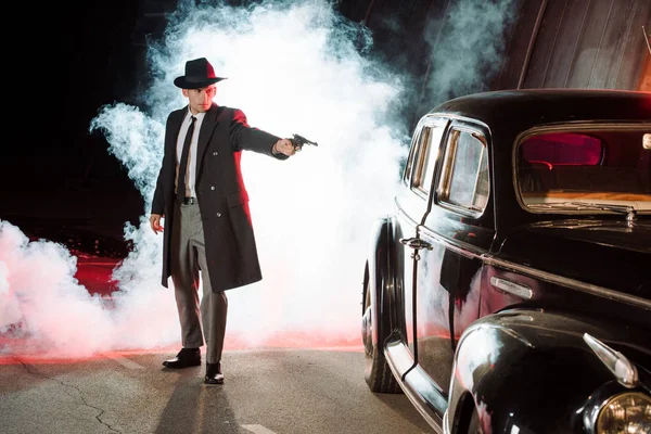 Gangster elegante em chapéu segurando arma perto de carro retro e fumaça — Fotografia de Stock