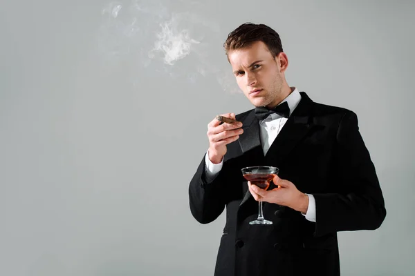 Багатий чоловік у костюмі з краваткою, що тримає келих з алкогольним напоєм та сигарою ізольовано на сірому — стокове фото