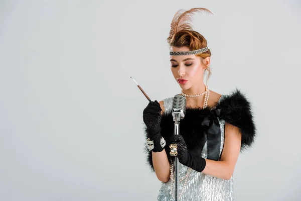 Attractive woman touching retro microphone and holding cigarette holder isolated on grey — Stock Photo