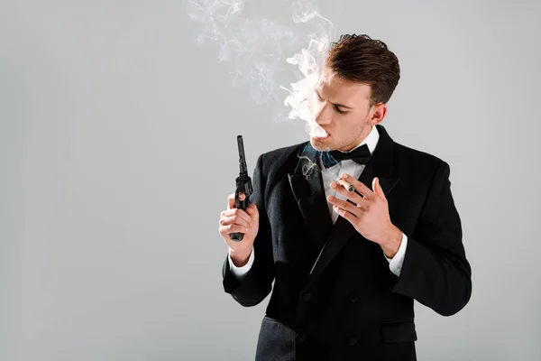 Hombre peligroso en traje con corbata de lazo sosteniendo arma mientras fuma cigarro aislado en gris - foto de stock