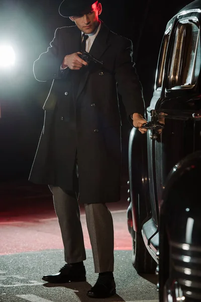 Stylish gangster in hat and coat holding gun near retro black car — Stock Photo
