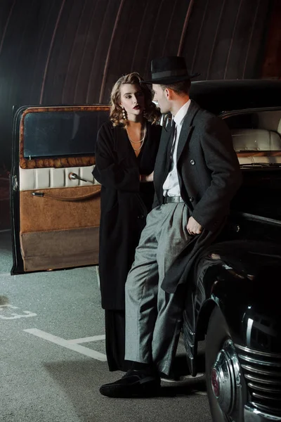 Handsome gangster in hat looking at attractive woman near retro car — Stock Photo