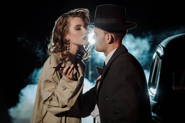 Handsome gangster in hat looking at attractive woman near car on black with smoke — Stock Photo