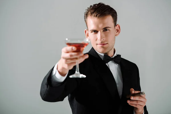 Enfoque selectivo del hombre guapo en traje con corbata de lazo celebración de vidrio con bebida y cigarro aislado en gris - foto de stock