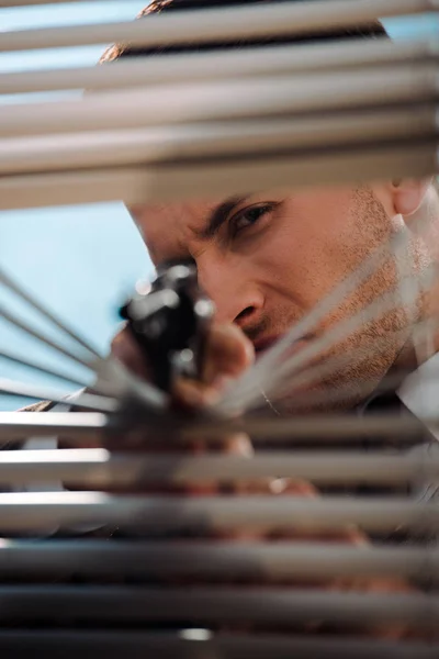 Вибірковий фокус небезпечної людини, що тримає пістолет біля віконних жалюзі — стокове фото