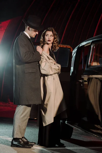 Hombre guapo en sombrero con abrigo en la mujer atractiva cerca de coche retro - foto de stock