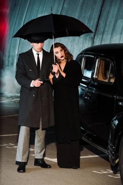 Handsome gangsters standing under umbrella near retro car outside — Stock Photo