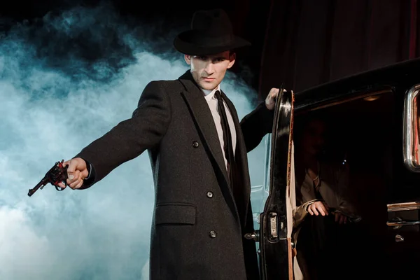 Dangerous man in hat holding gun near retro car on black with smoke — Stock Photo