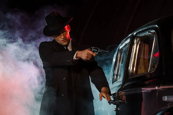 Stylish gangster holding gun near retro car on black with smoke — Stock Photo