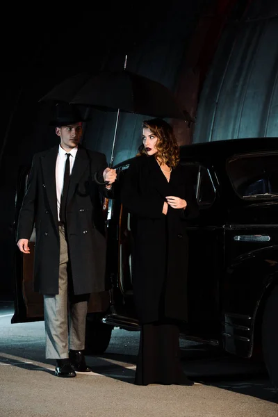 Handsome gangster holding umbrella near woman and retro car — Stock Photo