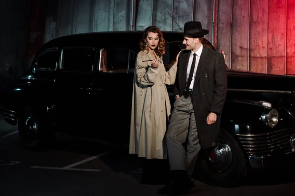 Glücklicher Gangster mit Hut sieht Frau mit Zigarette in der Nähe von Retro-Auto an — Stockfoto