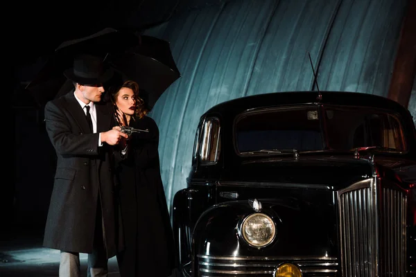 Небезпечний чоловік в капелюсі тримає пістолет і парасольку біля жінки і ретро автомобіль — стокове фото