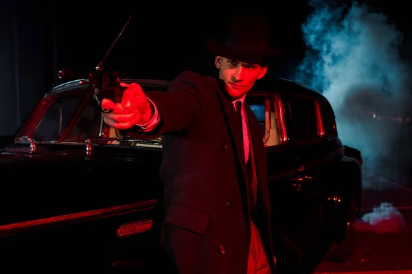 Selective focus of stylish man in hat holding gun near retro car on black with smoke — Stock Photo