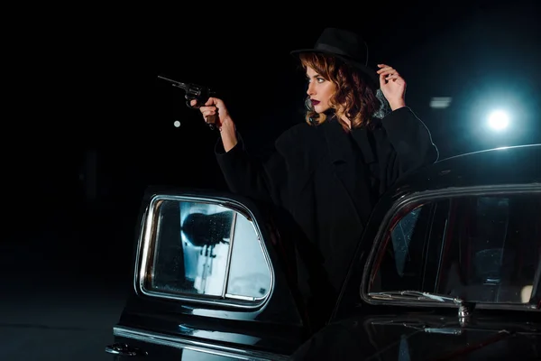 Dangerous woman in hat holding gun near retro car on black — Stock Photo