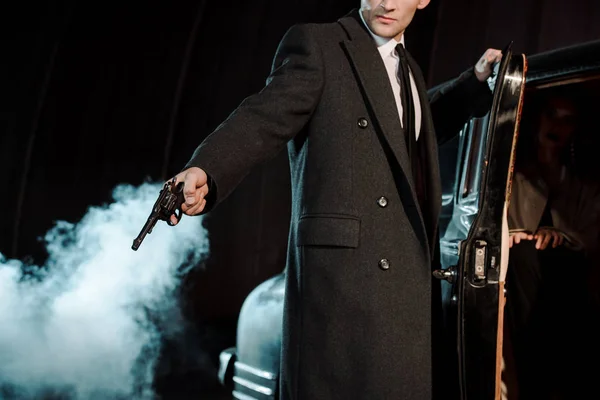 Cropped view of gangster holding gun near woman in vintage car on black with smoke — Stock Photo