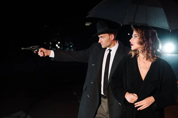 Vista lateral de hombre peligroso en sombrero sosteniendo pistola cerca de chica atractiva y coche retro en negro - foto de stock