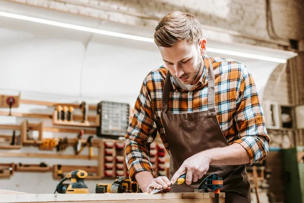 Bearded carpenter holding chisel while carving wood in workshop — Stock Photo