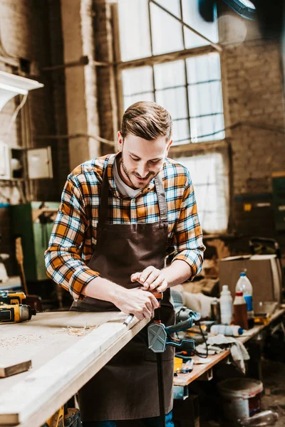 Selective focus of handsome woodworker holding chisel while carving wood in workshop — Stock Photo