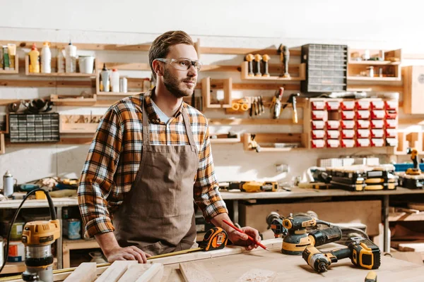 Carpinteiro bonito em óculos de pé perto da mesa e do equipamento — Fotografia de Stock