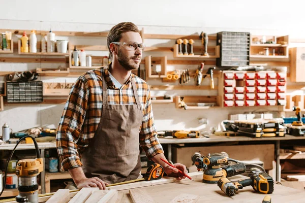 Beau menuisier dans des lunettes debout près de la table et de l'équipement — Photo de stock