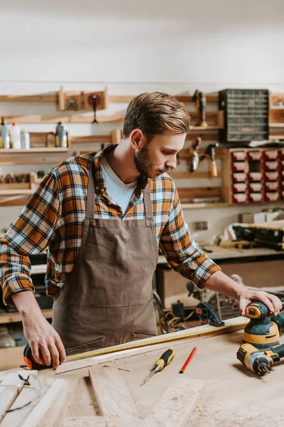 Beau menuisier en tablier brun mesurant la planche de bois — Photo de stock