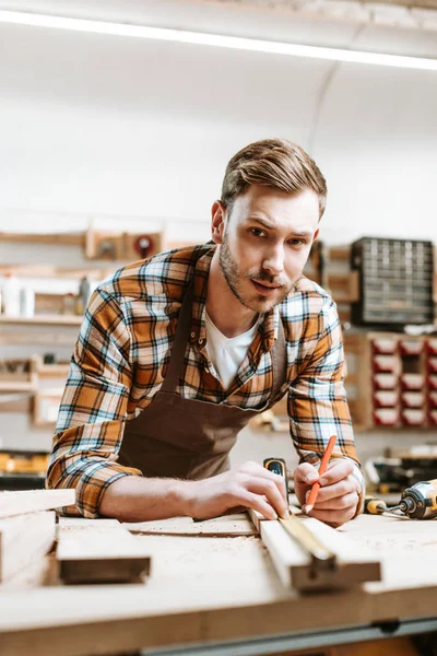 Selective focus of handsome woodworker holding pencil while measuring wooden plank — Stock Photo