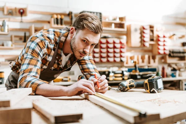Selektiver Fokus eines gut aussehenden Holzarbeiters, der Bleistift hält, während er Holzplanken misst — Stockfoto