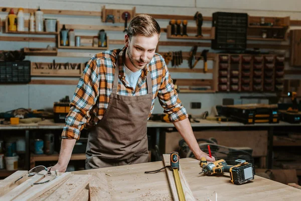 Handsome woodworker looking at table with wooden planks and measuring tape — Stock Photo