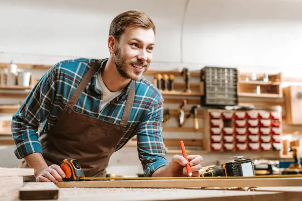 Selektiver Fokus eines fröhlichen bärtigen Holzarbeiters mit Bleistift in der Nähe von Maßband in der Werkstatt — Stockfoto