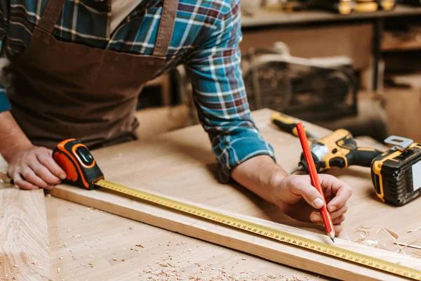 Cropped view of carpenter measuring plank with tape-measure in workshop — Stock Photo