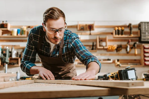 Selective focus of handsome carpenter in goggles touching wooden plank — Stock Photo