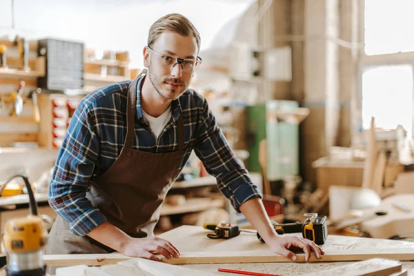 Selektiver Fokus des gutaussehenden Tischlers in Brille, der neben Holzplanke auf dem Tisch steht — Stockfoto