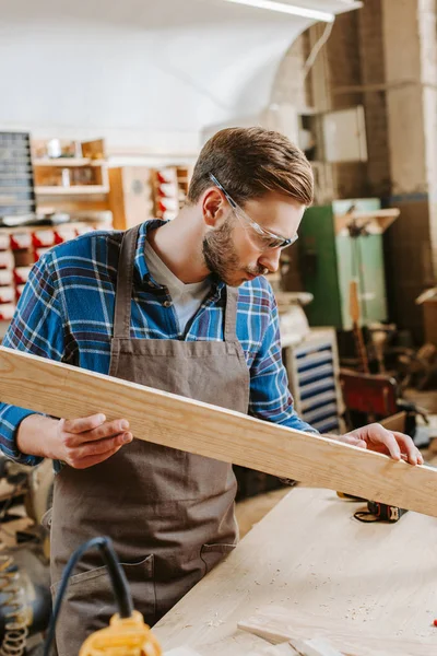 Selective focus of handsome carpenter in goggles holding wooden plank in workshop — Stock Photo