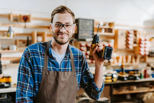 Happy carpenter in goggles and apron holding hammer drill — Stock Photo
