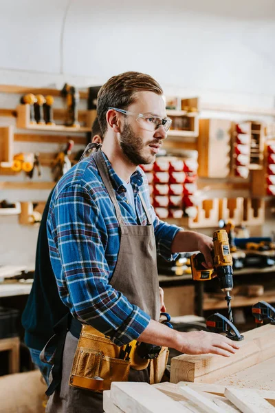 Tischler mit Schutzbrille und Schürze hält Hammerbohrer in der Nähe von Holzplanken — Stockfoto