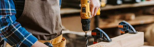 Plano panorámico de carpintero en delantal sosteniendo taladro de martillo cerca de tablón de madera — Stock Photo