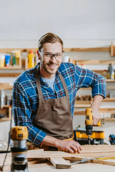 Selective focus of cheerful carpenter in goggles and apron holding hammer drill near wooden planks — Stock Photo